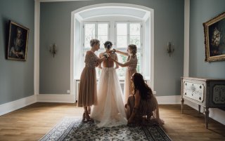 Eine Braut in einem wunderschönen Kleid steht vor einem Fenster und bereitet sich auf ihre Hochzeit vor, während ihre Brautjungfern ihr helfen.